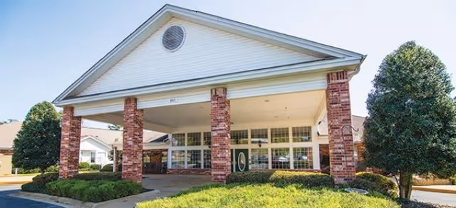 Exterior front view of a single-story building with a large covered entrance supported by brick columns, surrounded by green shrubs and trees under a clear sky.