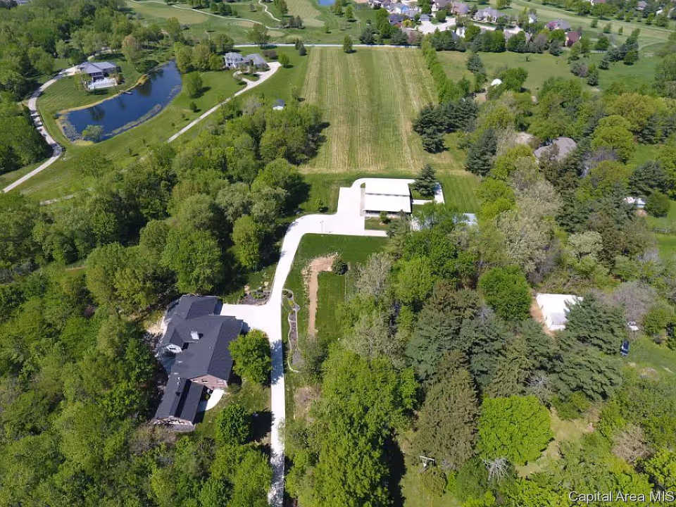 Aerial view of a senior living facility named Springfield Homes The Ranch, showing a large house surrounded by dense green trees, a winding driveway, a pond, open grassy fields, and nearby residential houses in the background.