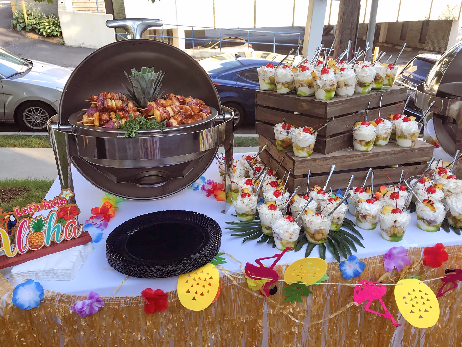 Outdoor buffet table decorated with a tropical theme including colorful flower garlands and paper cutouts of pineapples and flamingos. The table holds a large round chafing dish filled with skewers of grilled meat and vegetables, and multiple small dessert cups topped with whipped cream and cherries arranged on wooden crates. Black disposable plates and napkins are also on the table. Cars and a sidewalk are visible in the background.