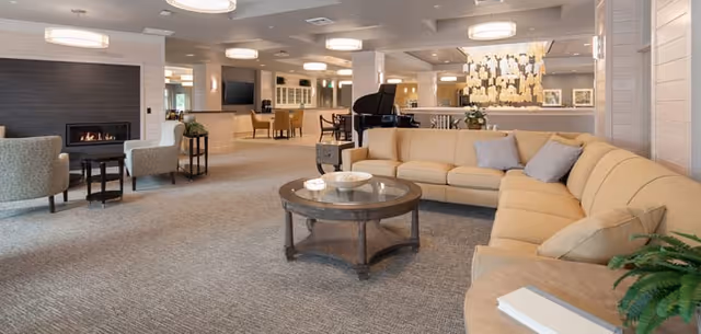 Spacious and well-lit senior living common area featuring a large beige sectional sofa with pillows, a round glass coffee table, a fireplace with two armchairs, a grand piano, and multiple seating areas with tables and chairs. The room has neutral tones, modern light fixtures, and decorative wall art.