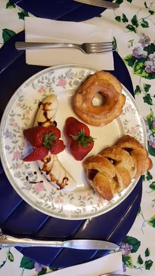 A floral plate holding assorted pastries, bananas drizzled with sauce, and strawberries on a floral tablecloth with a fork and napkin nearby.
