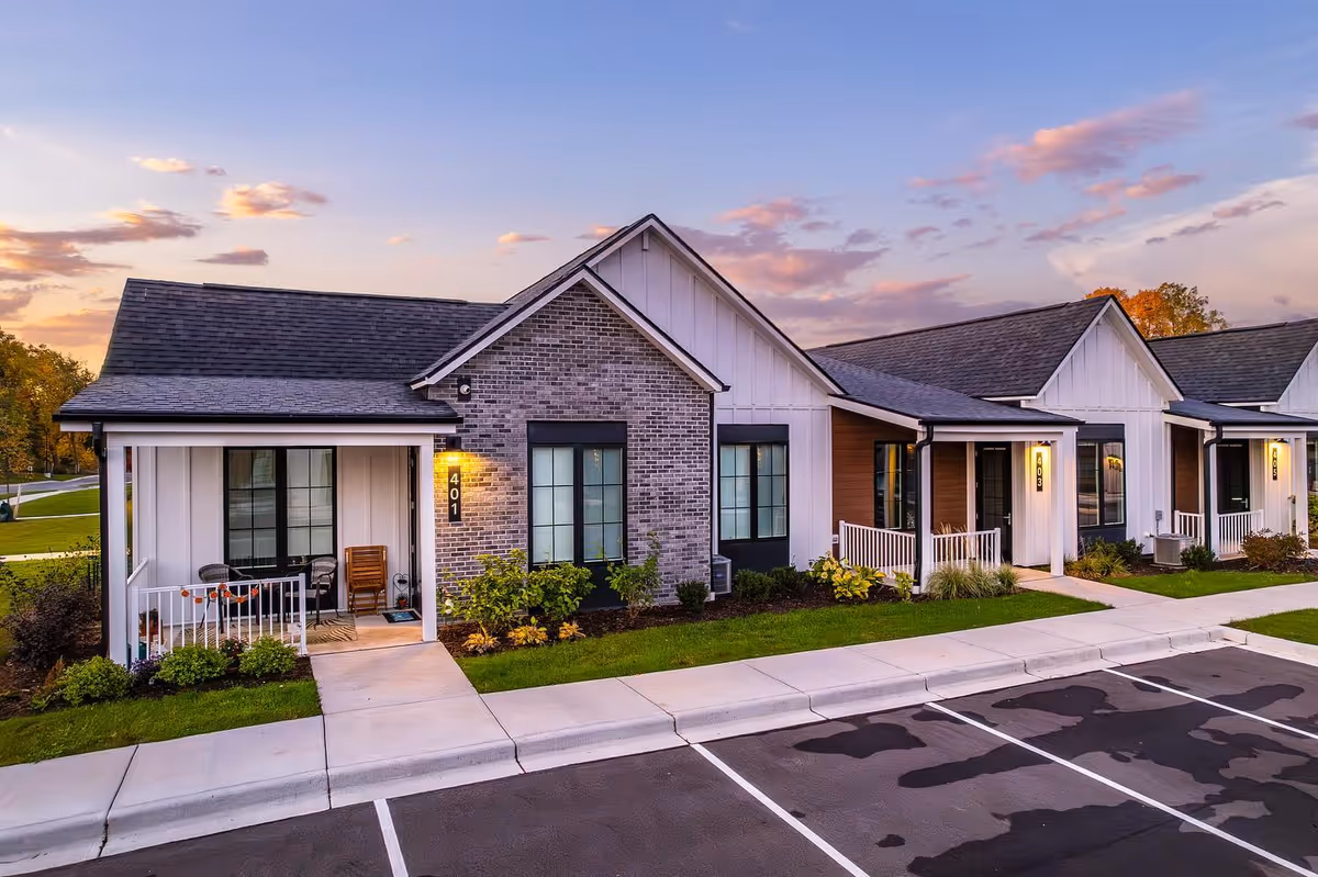 Exterior view of a single-story residential building at sunset with multiple units, each having a small porch with chairs and plants. The building features a combination of brick and siding with gabled roofs. There is a parking lot in front with marked spaces and a sidewalk leading to the entrances.