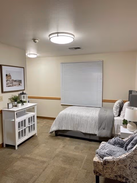 A cozy bedroom with a single bed covered in gray and white bedding, a patterned armchair with a gray throw blanket, a white cabinet with decorative items including a small plant and a lantern, a framed picture on the wall, and a window with closed blinds.