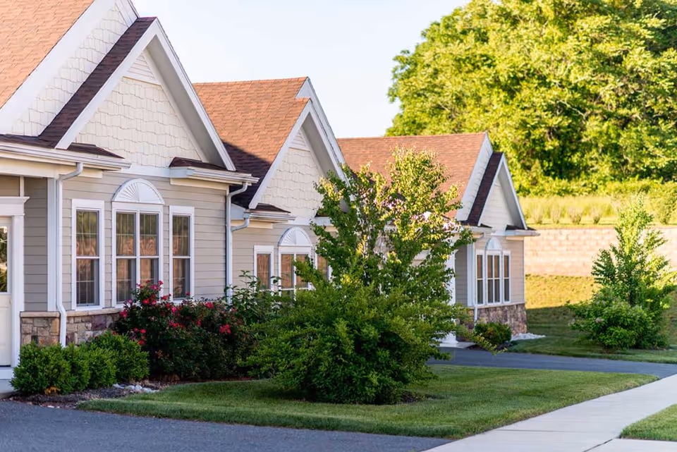Exterior view of a senior living facility with beige siding, white trim, and brown shingled roofs. The building is surrounded by well-maintained green lawns, bushes, and trees under a clear sky.