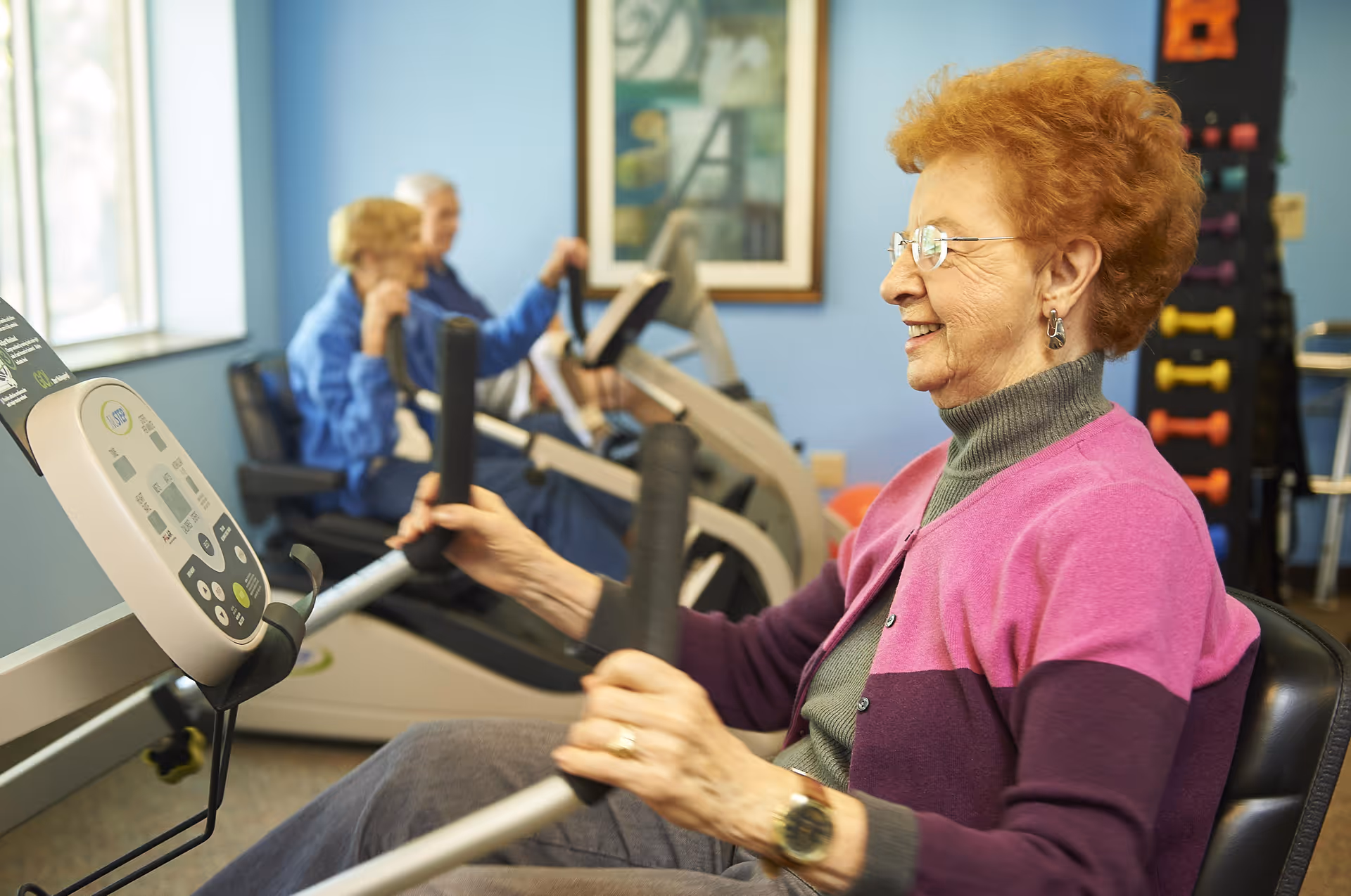 Three elderly women exercising on stationary recumbent bikes in a fitness room with blue walls and a window letting in natural light. One woman in the foreground is smiling and wearing glasses, a pink and purple cardigan, and a gray turtleneck.
