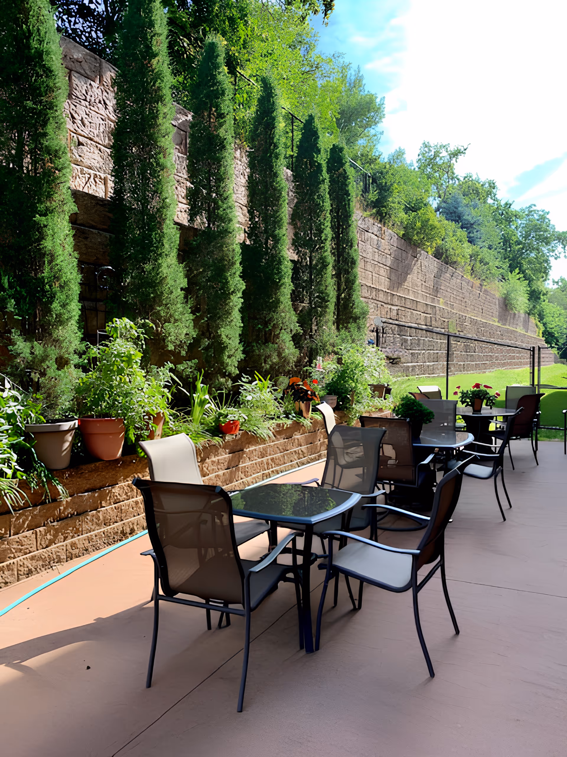 Outdoor patio area with several glass-top tables and mesh chairs arranged on a concrete surface. Alongside the patio is a raised garden bed with potted plants and tall green shrubs against a stone retaining wall. Trees and greenery are visible in the background under a partly cloudy sky.