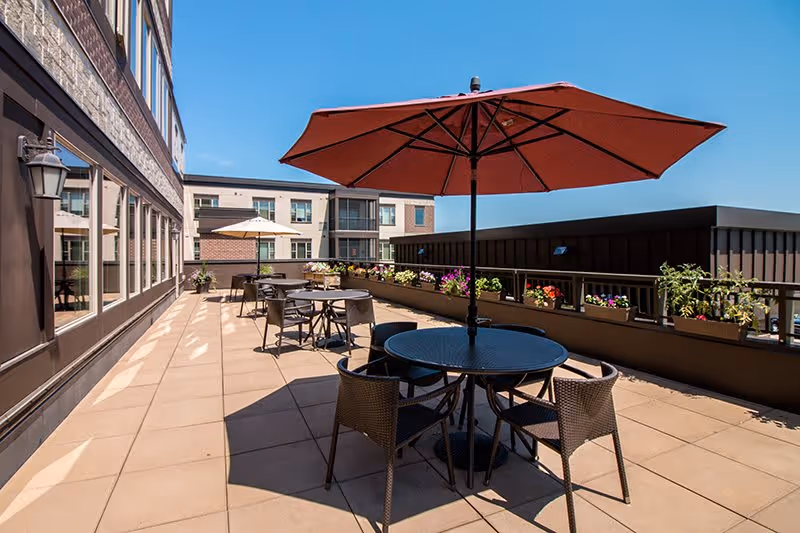 Outdoor patio area with multiple round tables and chairs, each table shaded by large umbrellas. Flower boxes with colorful flowers line the railing, and a multi-story building is visible in the background under a clear blue sky.