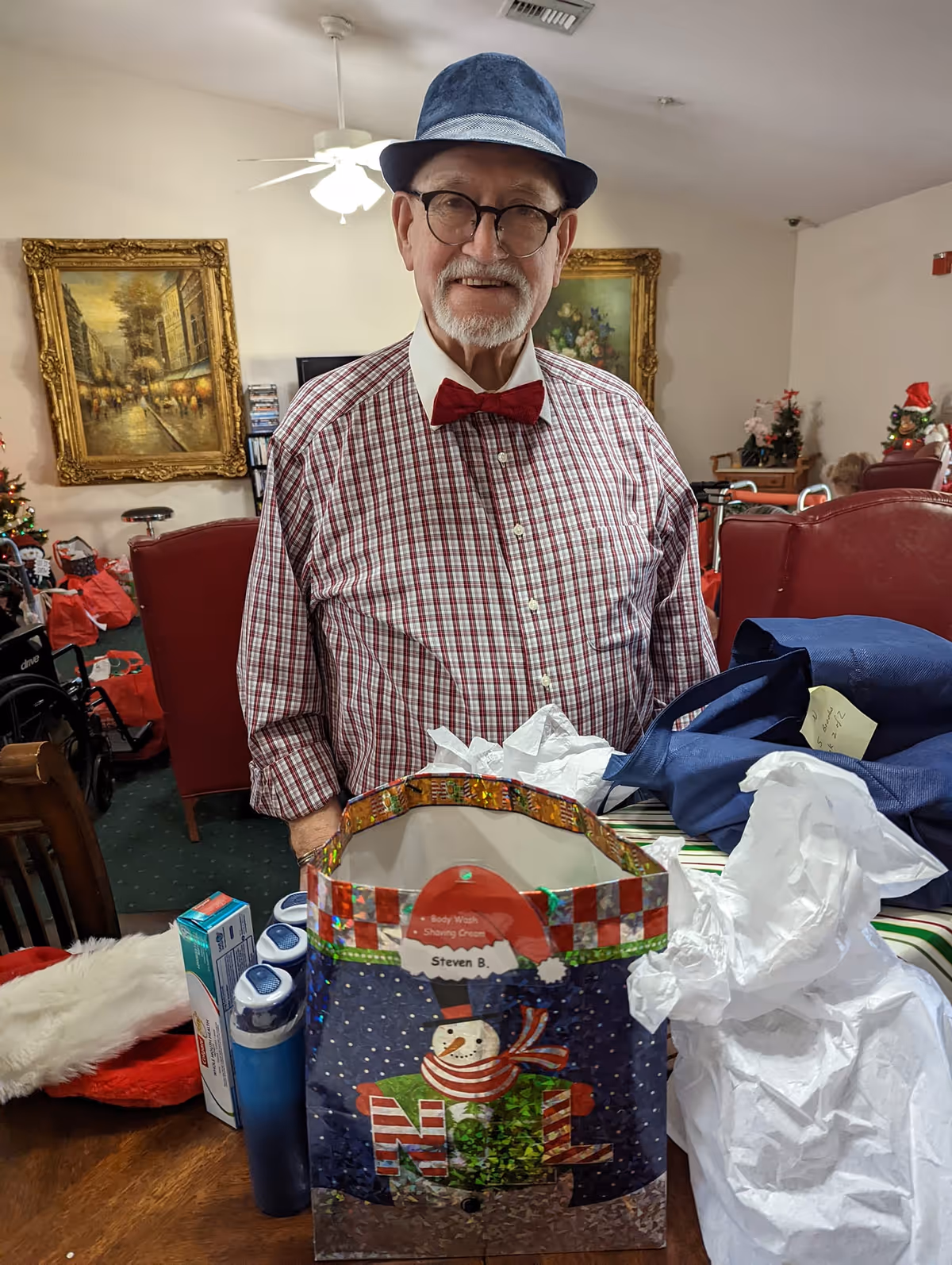 An elderly man wearing a blue hat, glasses, a red bow tie, and a checkered shirt stands indoors in a decorated room. In front of him on a table is a festive gift bag with a snowman design and some tissue paper, along with other items including a toothbrush and toothpaste. The room has framed paintings on the wall and red chairs, with holiday decorations visible in the background.