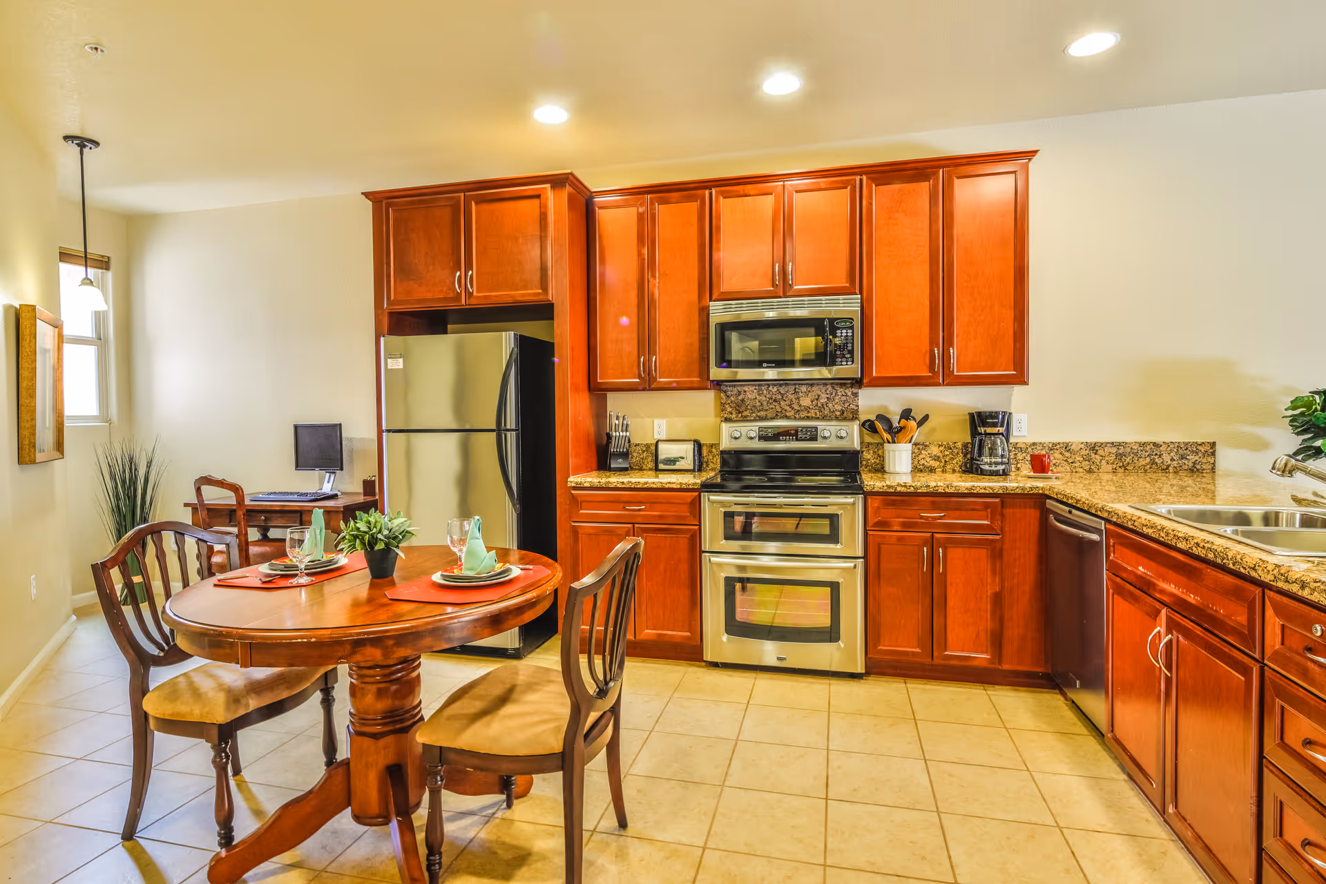 A bright kitchen with wooden cabinets, stainless steel refrigerator, oven, microwave, and dishwasher. There is a round wooden dining table set with two place settings and chairs. A small desk with a computer is visible in the background near a window.