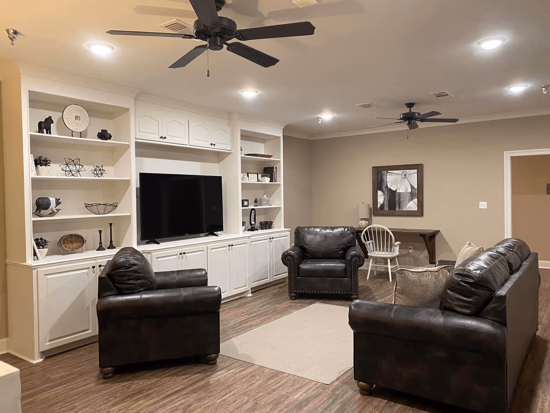 A cozy living room with leather sofas and armchairs facing a TV set in white built-in shelving and ceiling fans overhead.
