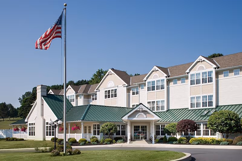 Exterior view of Mifflin Court, a multi-story senior living facility with beige siding and a green metal roof. The building has multiple large windows and a covered entrance. An American flag on a flagpole is prominently displayed in front of the building, with well-maintained landscaping including bushes and flowers surrounding the entrance.