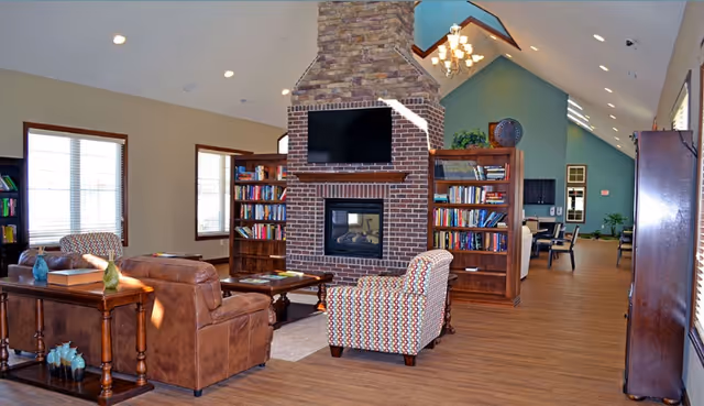 A spacious living room area with a high vaulted ceiling, featuring a central brick fireplace with a mounted flat-screen TV above it. Surrounding the fireplace are wooden bookshelves filled with books. The room has comfortable seating including a brown leather sofa and patterned armchairs, with wooden tables and decorative items. Large windows allow natural light to fill the space, and the flooring is wood. The room extends into a dining or common area in the background.
