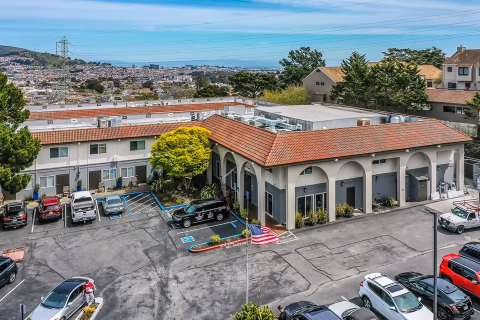 Aerial view of Serra Highlands Senior Living facility showing a building with a red-tiled roof, parking lot with several parked cars, an American flag on a flagpole, and surrounding trees and residential area in the background.