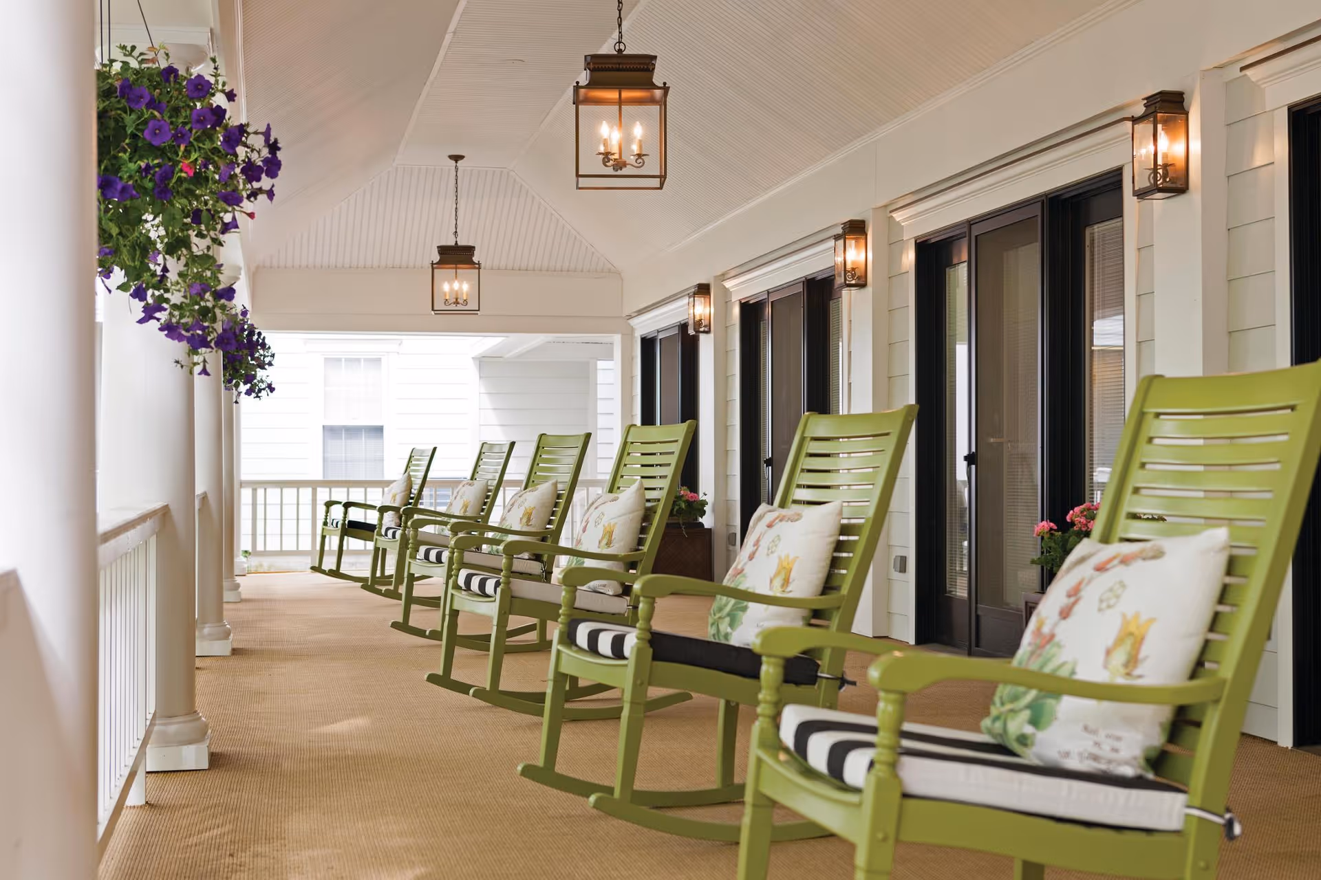 Covered porch with a row of green rocking chairs with patterned cushions, hanging lanterns, and potted flowers.