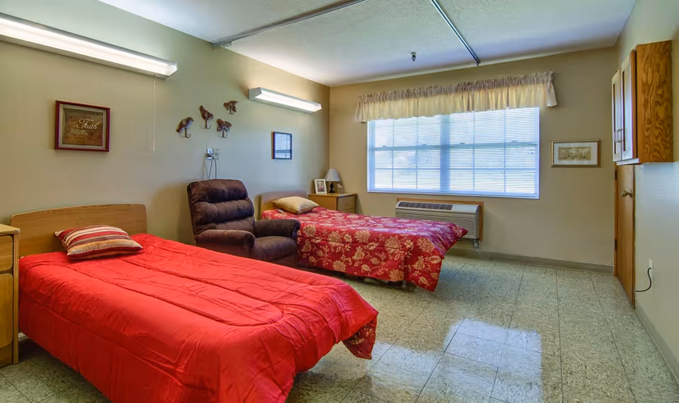 A simple, clean bedroom with two single beds, one with a red bedspread and striped pillow, the other with a floral red bedspread and beige pillow. Between the beds is a brown recliner chair. The room has a large window with blinds and a valance, a small nightstand with a lamp and picture frame, wall decorations including a framed picture and three bird hooks, and a wooden cabinet on the right wall.