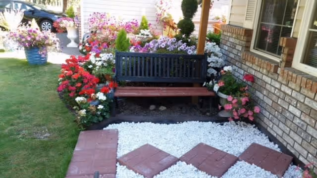 A garden area with a black and brown wooden bench surrounded by colorful flowers including red, pink, white, and purple blooms. The ground in front of the bench is covered with white gravel and red square stepping stones arranged in a diamond pattern. The garden is adjacent to a brick building with windows.