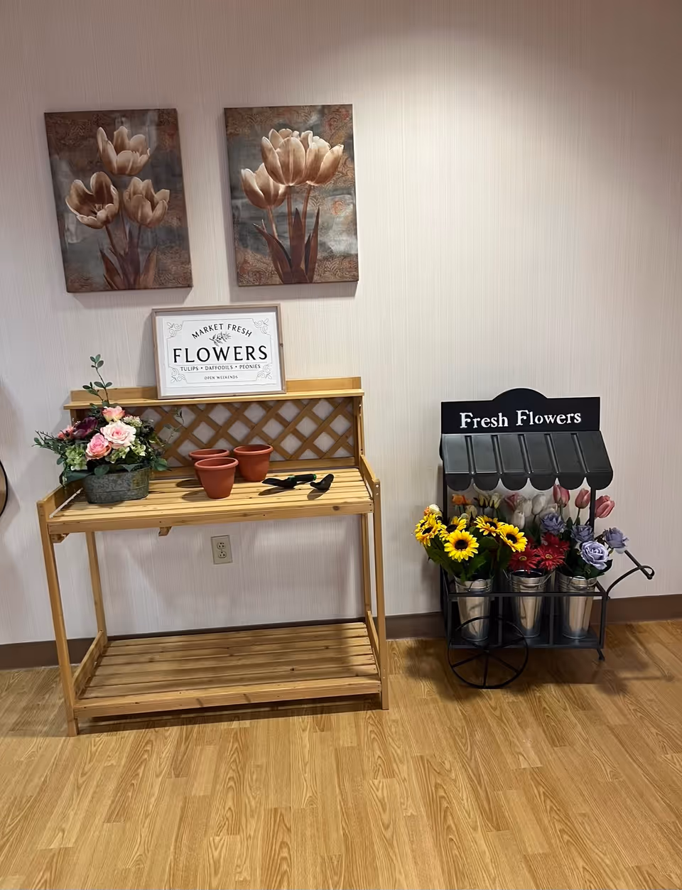 A wooden potting bench and a small flower cart labeled "Fresh Flowers" with artificial blooms beneath two floral wall paintings on a wood floor.