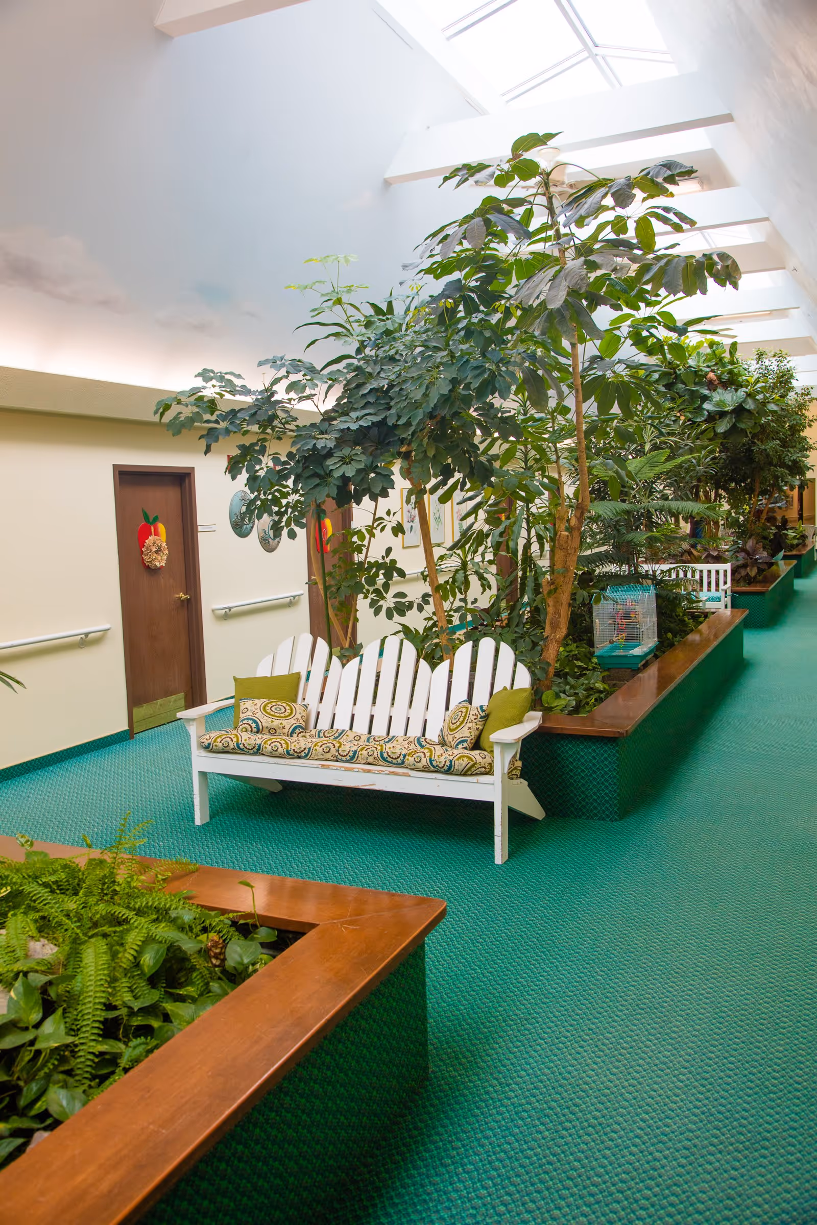 Indoor hallway with green carpet and large planters filled with various green plants. White wooden bench with patterned cushions is placed next to the planters. Skylights above provide natural light. Doors with apple decorations are visible along the wall.
