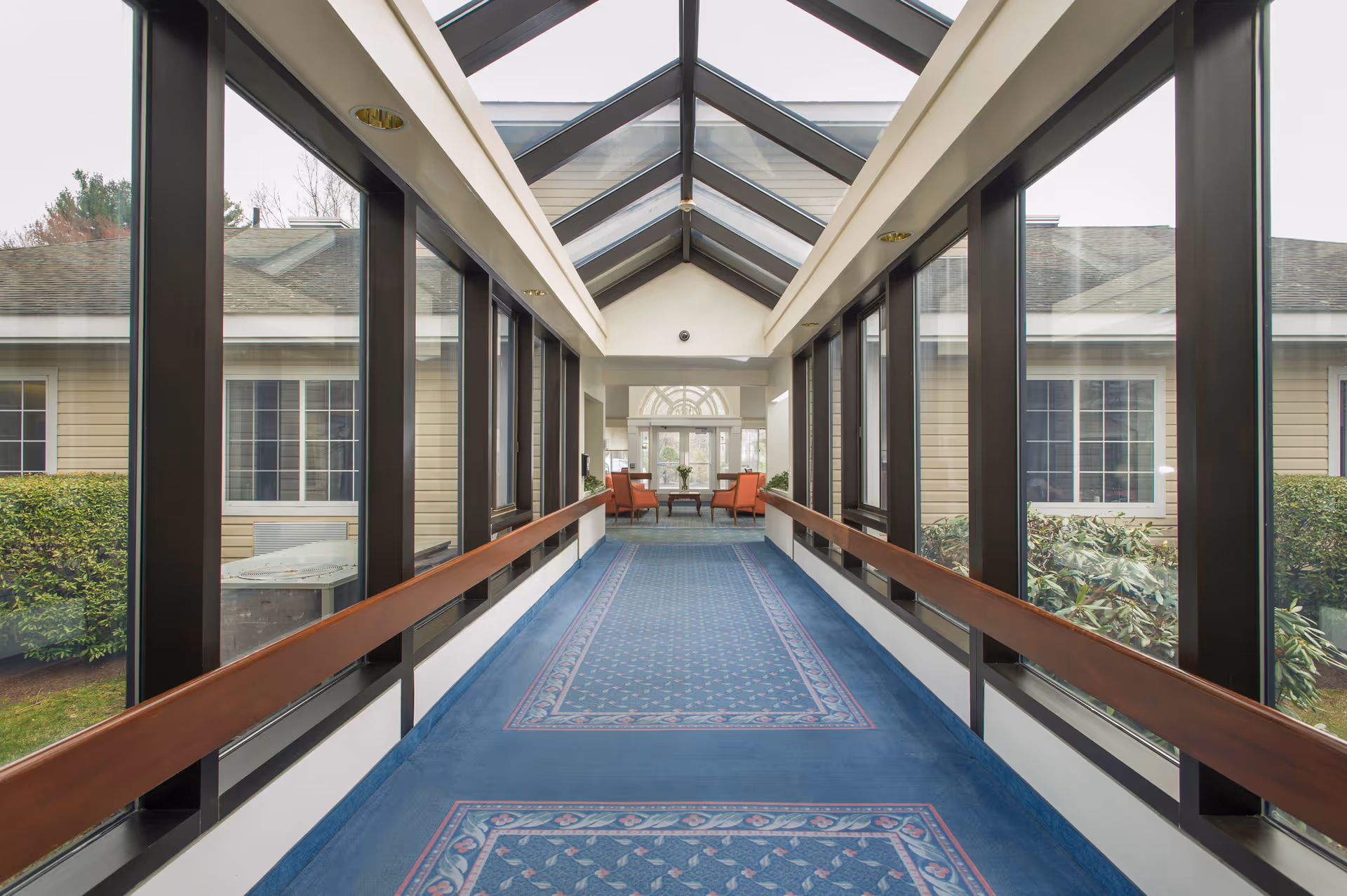 A long indoor corridor with large windows on both sides and a glass ceiling. The corridor has blue patterned carpet and wooden handrails. At the end of the corridor, there are two orange chairs and a small table with a flower vase, with a view of an exterior door and windows.