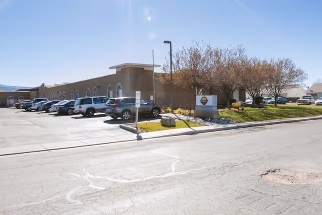 Exterior view of Cedar Health and Rehabilitation facility showing a single-story building with several parked cars in the parking lot, trees with sparse leaves, a street lamp, and a sign near the entrance on a sunny day.