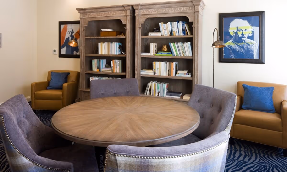 Round wooden table surrounded by upholstered chairs in a cozy common room with bookshelves and armchairs.