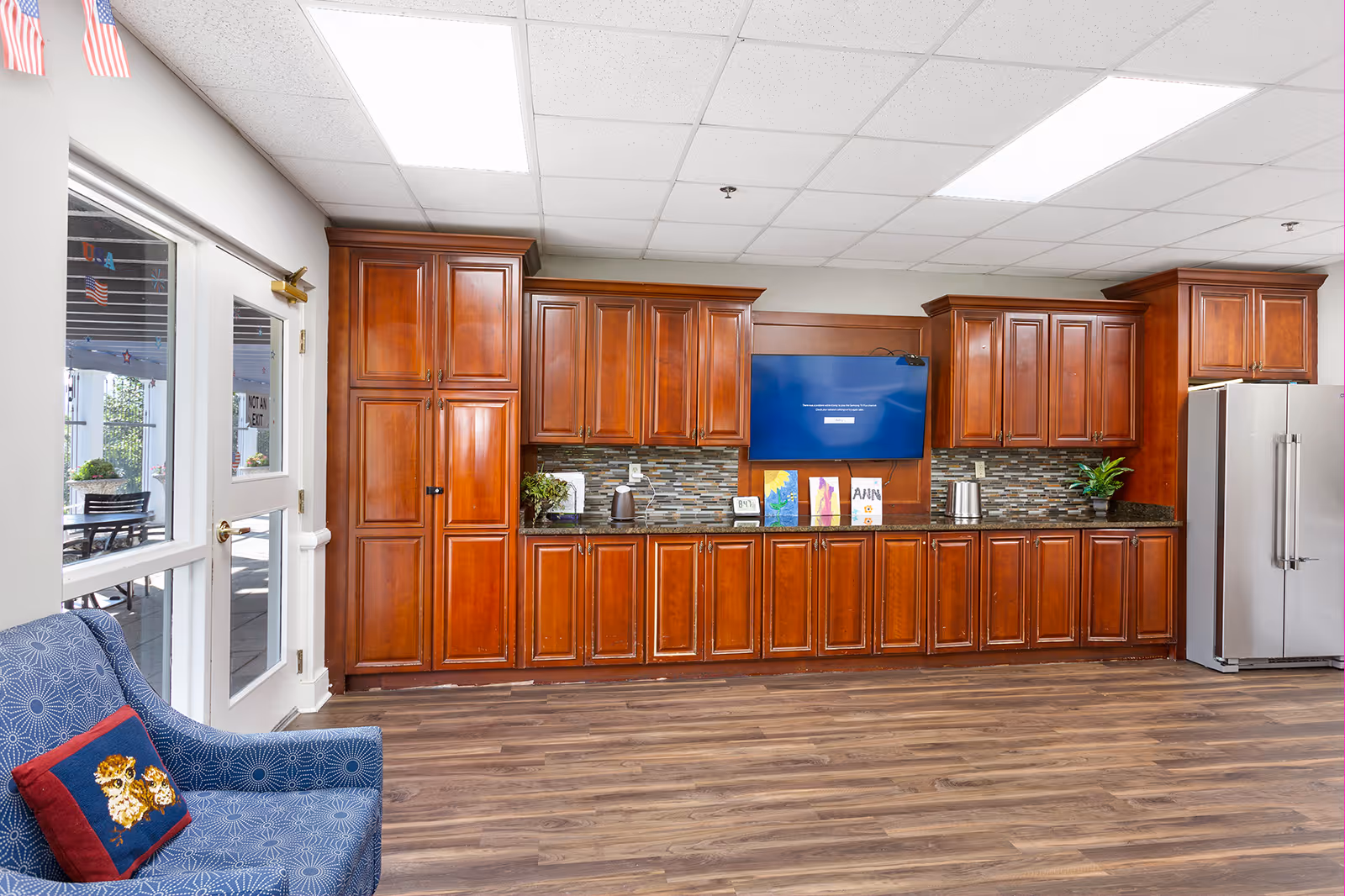 Interior view of a room with wooden cabinets along the wall, a mounted flat-screen TV, a stainless steel refrigerator, and a blue armchair with a decorative pillow featuring two owls. The room has a wood floor and a ceiling with recessed lighting. There is a glass door on the left side leading to an outdoor area with tables and chairs.