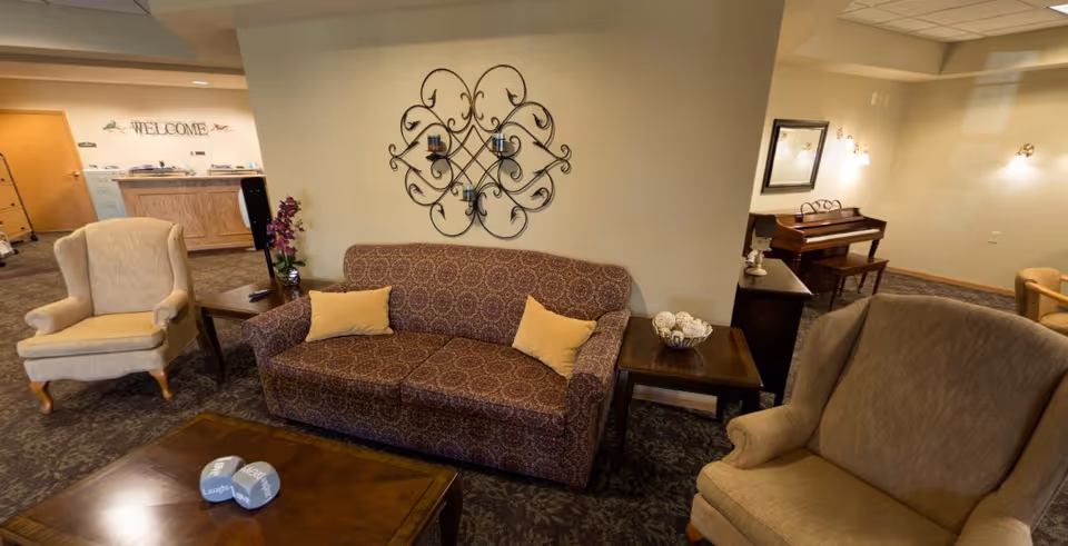 A cozy living room area with a patterned sofa and two beige armchairs surrounding a wooden coffee table. On the wall above the sofa is a decorative wrought iron candle holder. In the background, there is a piano with a bench, a mirror, and a welcome sign near a wooden counter.