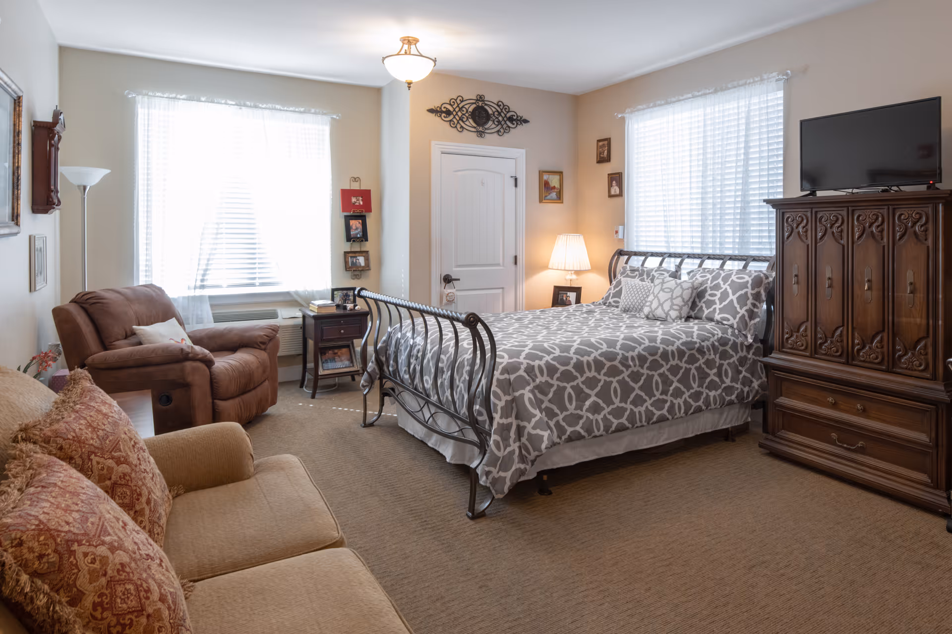 A cozy bedroom in an assisted living facility featuring a metal-framed bed with gray and white patterned bedding, a wooden dresser with a TV on top, a brown recliner chair, a beige sofa with patterned cushions, two windows with white blinds and sheer curtains, a nightstand with a lamp, and framed pictures on the walls.