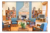 Communal lounge with upholstered armchairs arranged around a coffee table facing a stone fireplace, flanked by bookshelves, flags, and a TV.