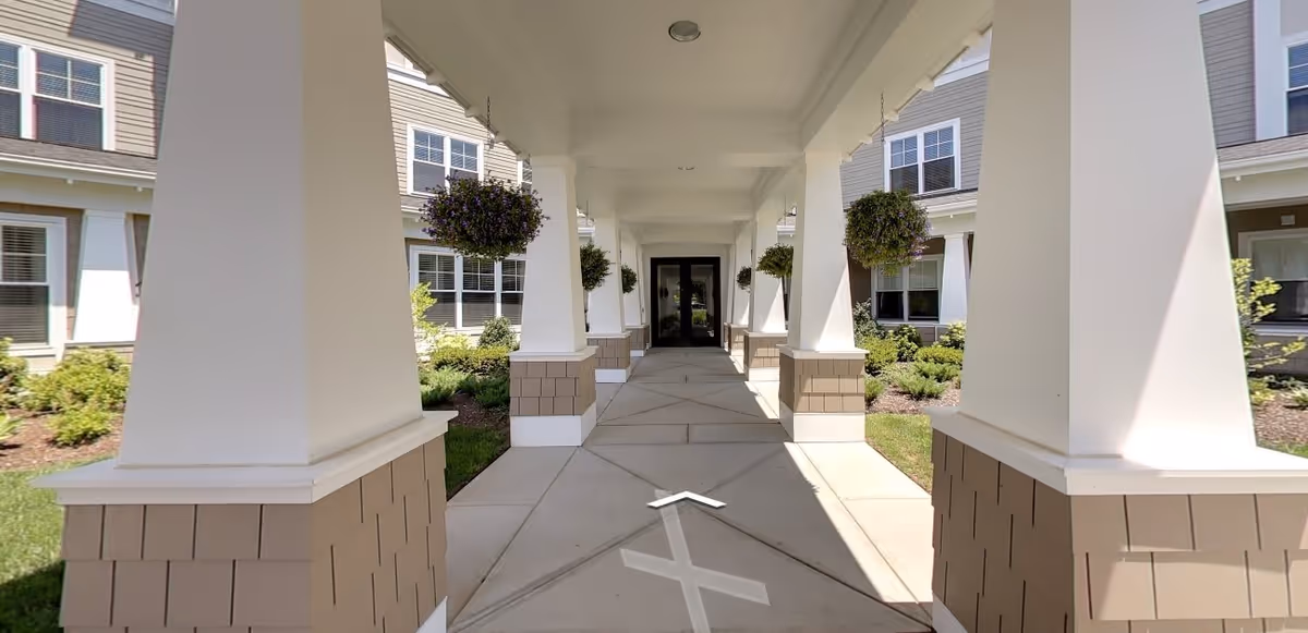 Covered walkway with large white columns and hanging flower baskets leading to a glass door entrance of a building, surrounded by landscaped greenery and windows.