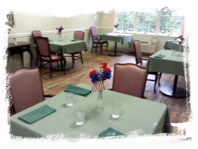 A dining room with several tables covered in green tablecloths, each set with green napkins, glasses of water, and small flower arrangements featuring red, white, and blue flowers with miniature American flags. The room has wooden flooring and large windows letting in natural light.