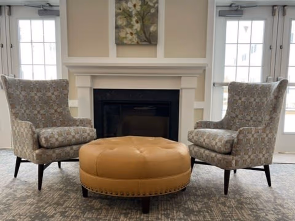 Two patterned armchairs facing each other with a round tan leather ottoman in between, positioned in front of a white fireplace with a floral painting above it. There are glass doors on either side of the fireplace letting in natural light.