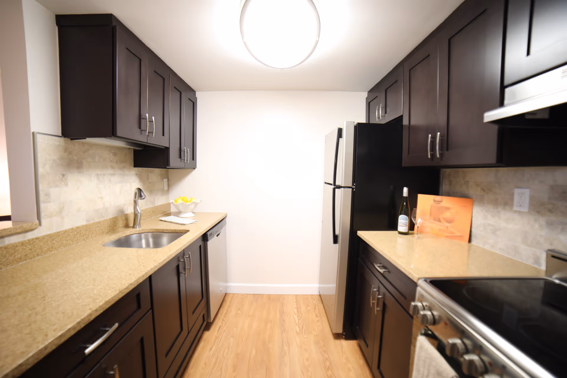 A modern kitchen with dark wood cabinets, beige countertops, a stainless steel sink, dishwasher, refrigerator, and stove. The floor is light wood, and there is a round ceiling light fixture. A bottle of wine and a wine glass are placed on the right countertop.