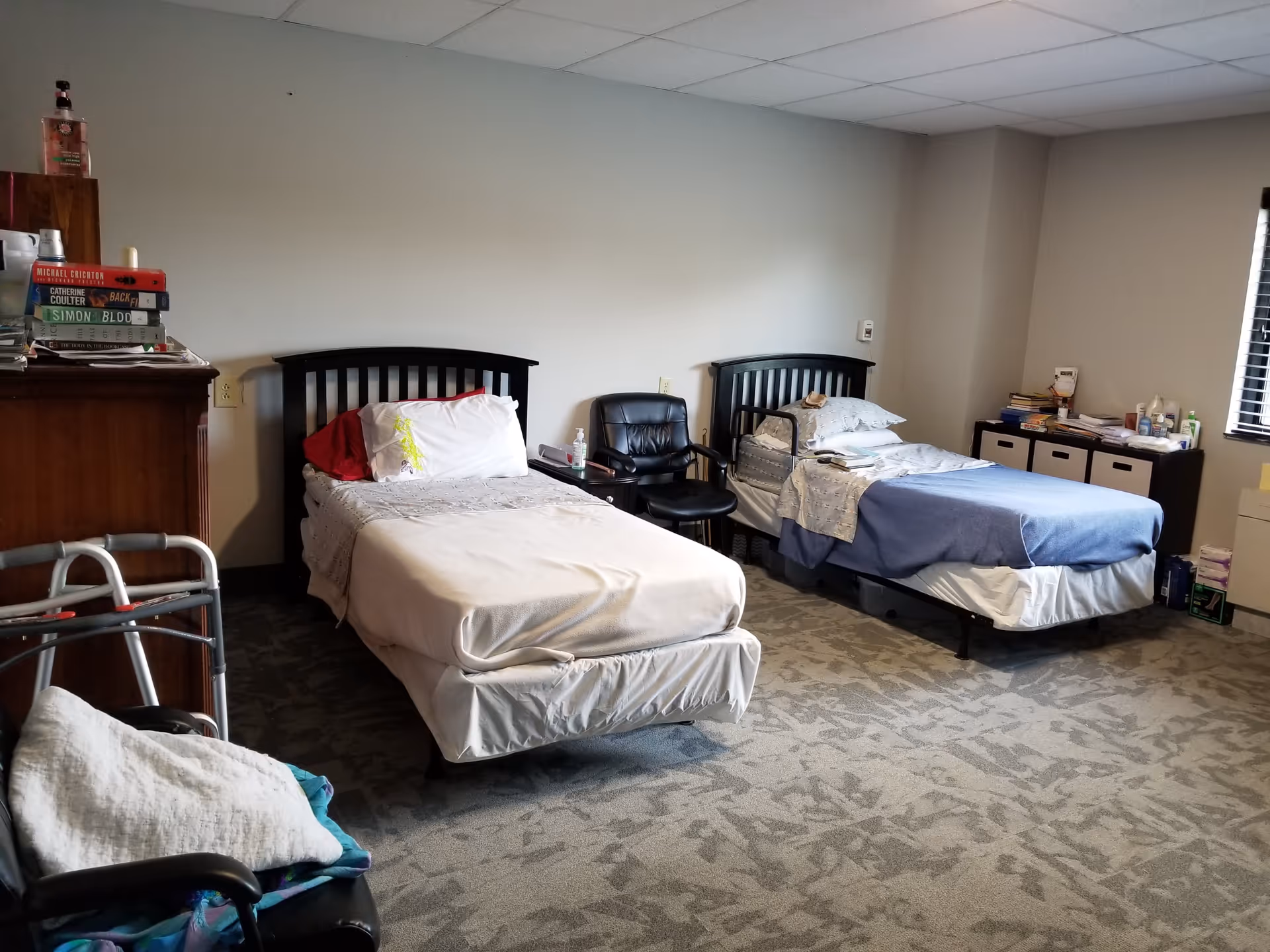 A bedroom with two single beds, each with a headboard and bedding. Between the beds is a black chair and a small table with a bottle of hand sanitizer. To the left, there is a wooden dresser with a stack of books and a bottle of hand soap on top. A walker is partially visible in the foreground. On the right side, there is a window with blinds and a cabinet with storage bins and various items on top. The room has a patterned carpet and a drop ceiling with fluorescent lighting.