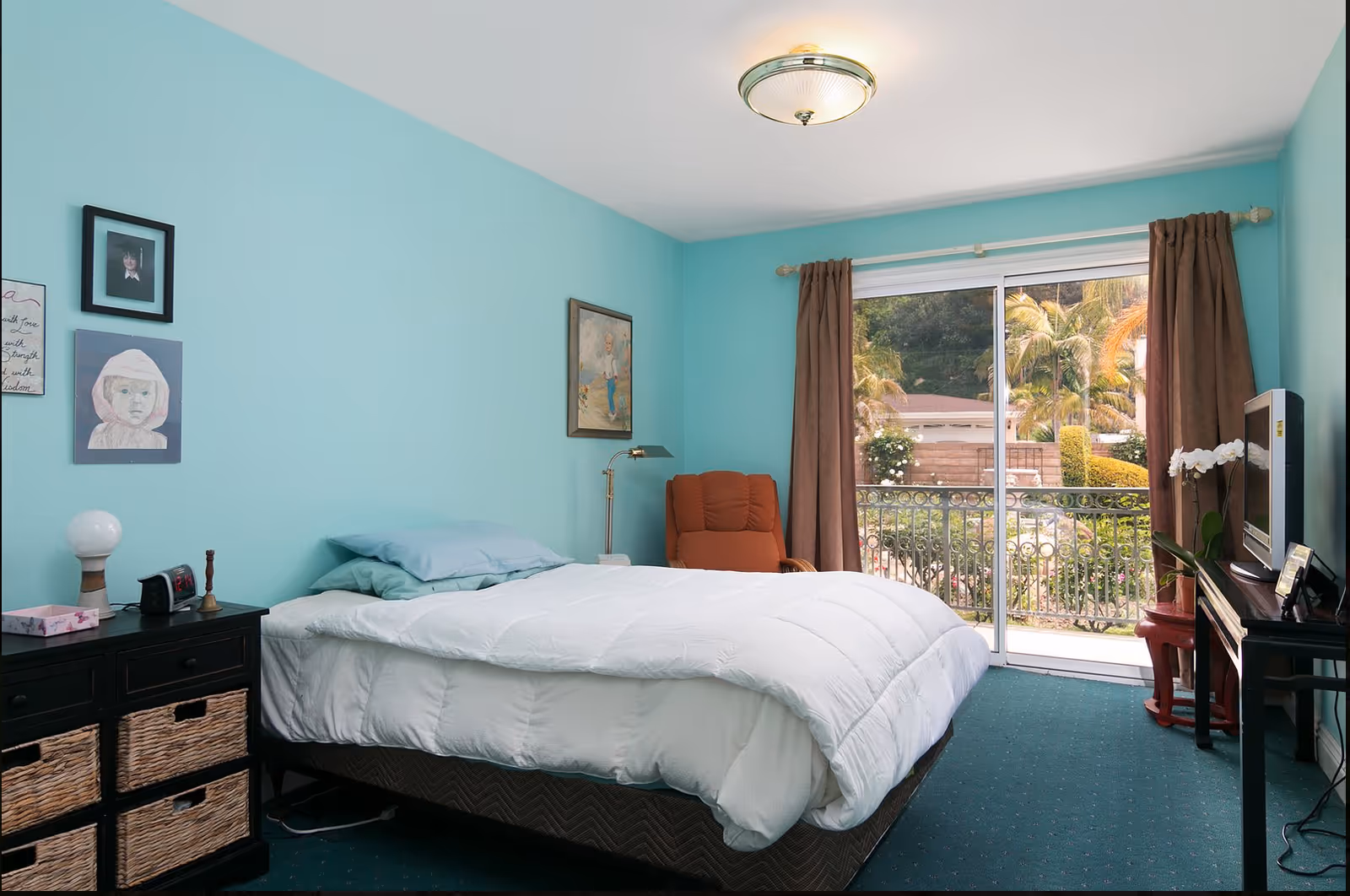 A bedroom with light blue walls featuring a bed with white bedding and blue pillows. There is a black nightstand with wicker drawers on the left side holding a lamp, clock, and other small items. A framed picture and artwork hang on the wall above the bed. A brown armchair is positioned near a sliding glass door with brown curtains, leading to an outdoor view with greenery and palm trees. A TV sits on a black stand to the right of the bed with a white orchid plant beside it.
