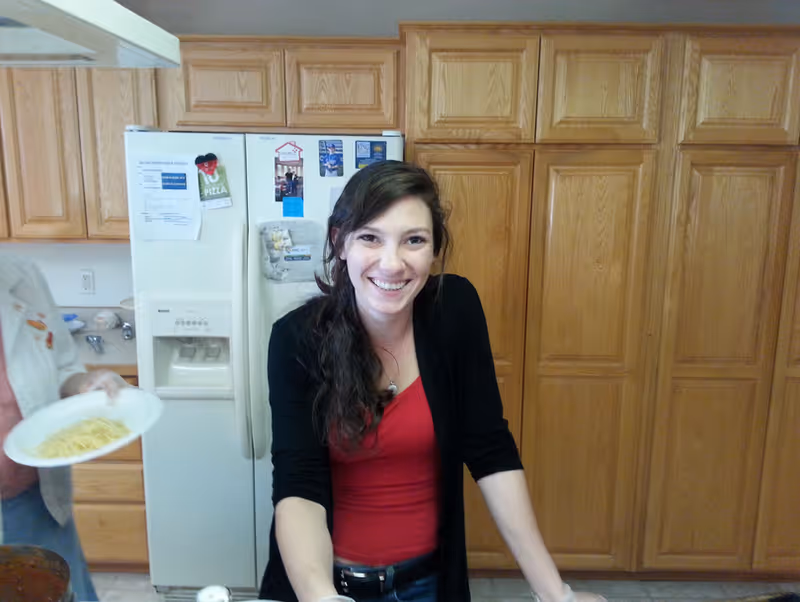 A smiling woman stands at a kitchen counter in front of a refrigerator and wooden cabinets.