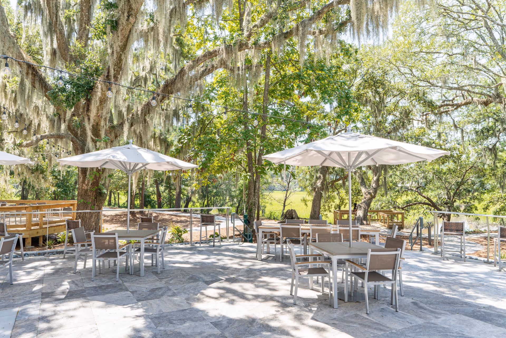 Outdoor patio with tables, chairs, and umbrellas beneath large oak trees draped with Spanish moss.