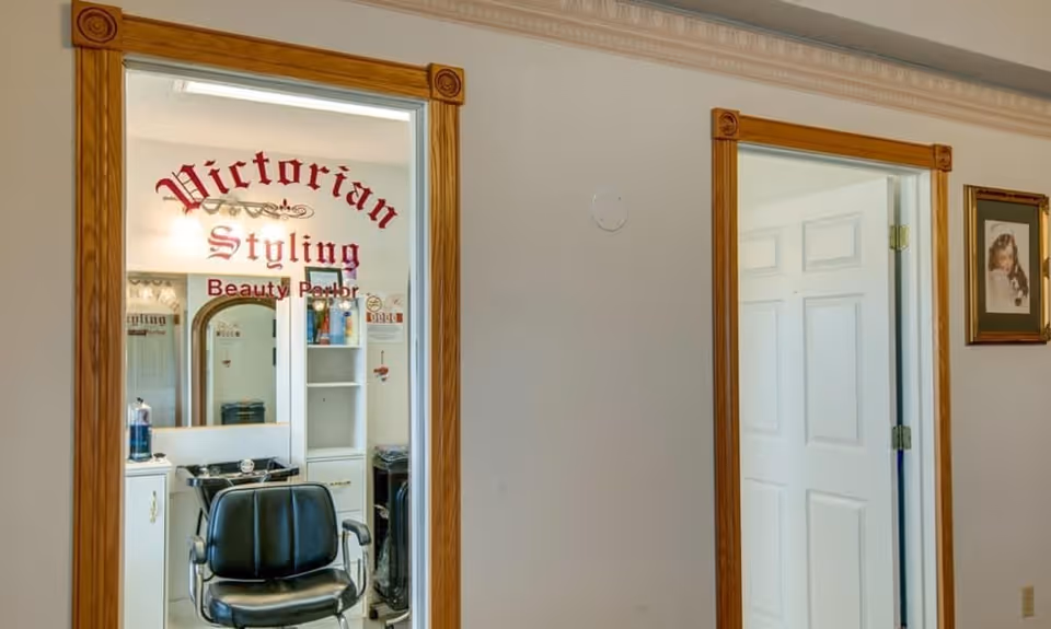 Hallway view into a small beauty parlor labeled "Victorian Styling" showing a black salon chair and cabinetry through a glass door.
