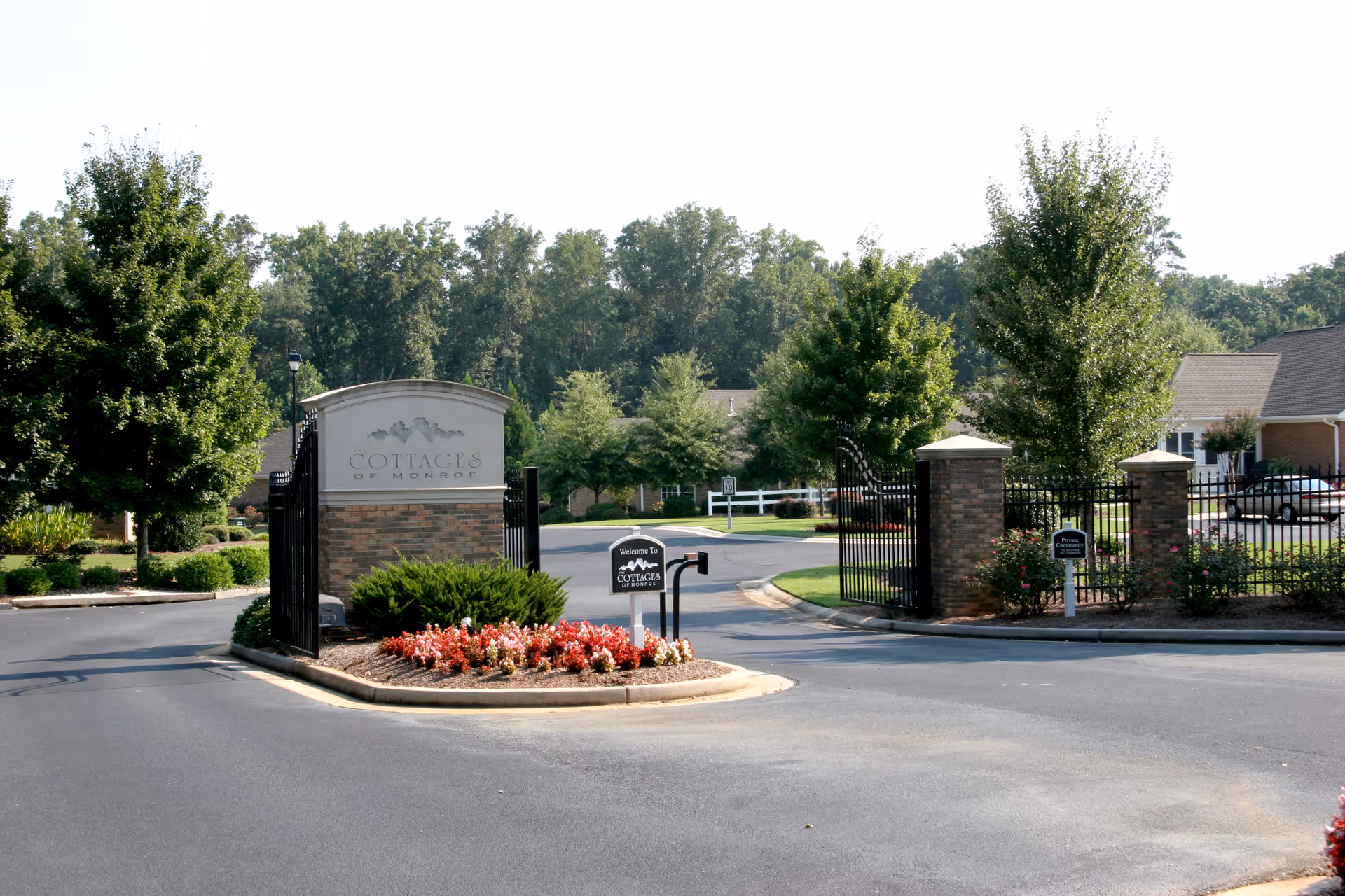 Entrance gate to The Cottages of Monroe senior living facility with brick pillars, black metal gates, landscaped flower beds, and trees lining the driveway.