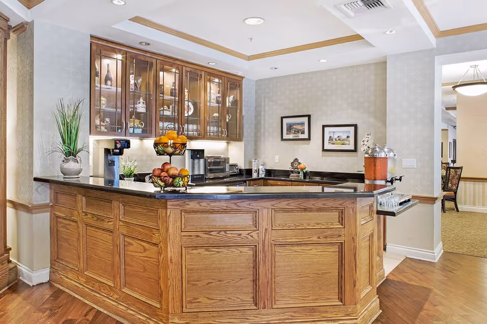 Wood-paneled communal kitchenette with a black countertop, glass-front cabinets, fruit displays, and a beverage station.