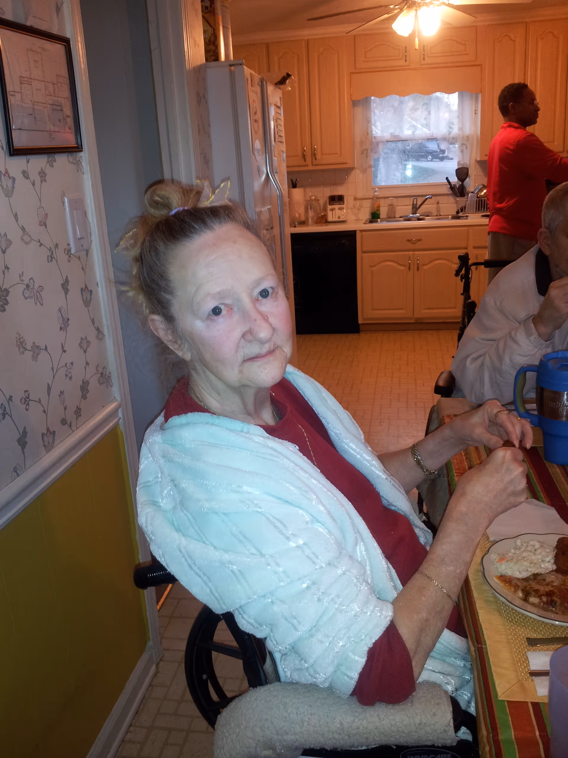 An elderly woman with light skin and blonde hair tied up in a bun sits in a wheelchair at a dining table with a plate of food in front of her. She is wearing a red shirt and a light blue shawl. In the background, a kitchen with wooden cabinets, a window with lace curtains, and a person in a red shirt standing near the sink are visible.