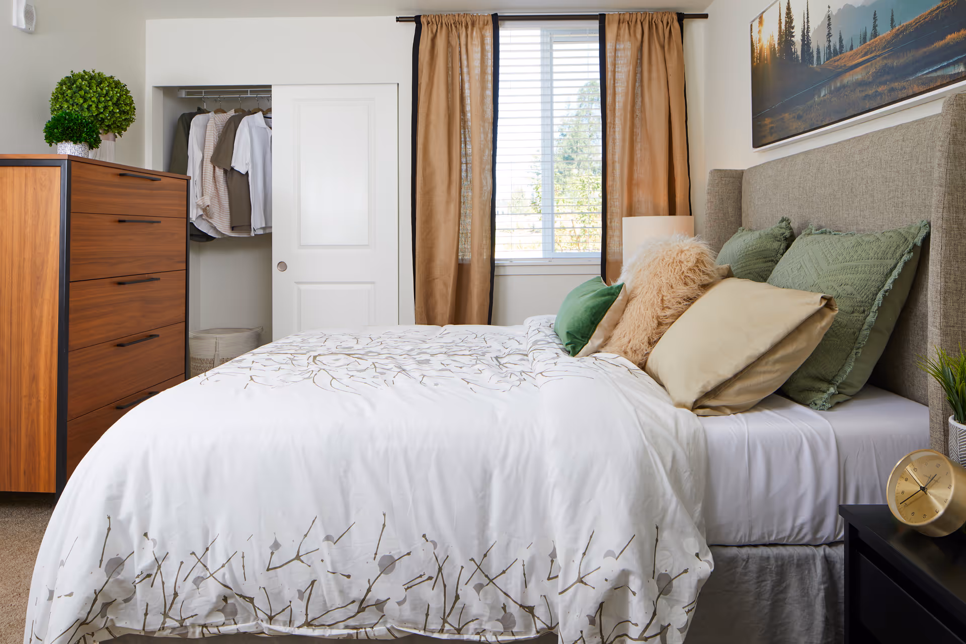 A neatly made bed with white and patterned bedding, several decorative pillows in green, beige, and fluffy textures, and a beige upholstered headboard. Next to the bed is a black nightstand with a gold clock and a small potted plant. On the left side of the room, there is a wooden dresser with a green potted plant on top and an open closet with clothes hanging inside. A window with beige curtains and white blinds is centered on the wall behind the bed, and a large landscape painting hangs above the headboard.