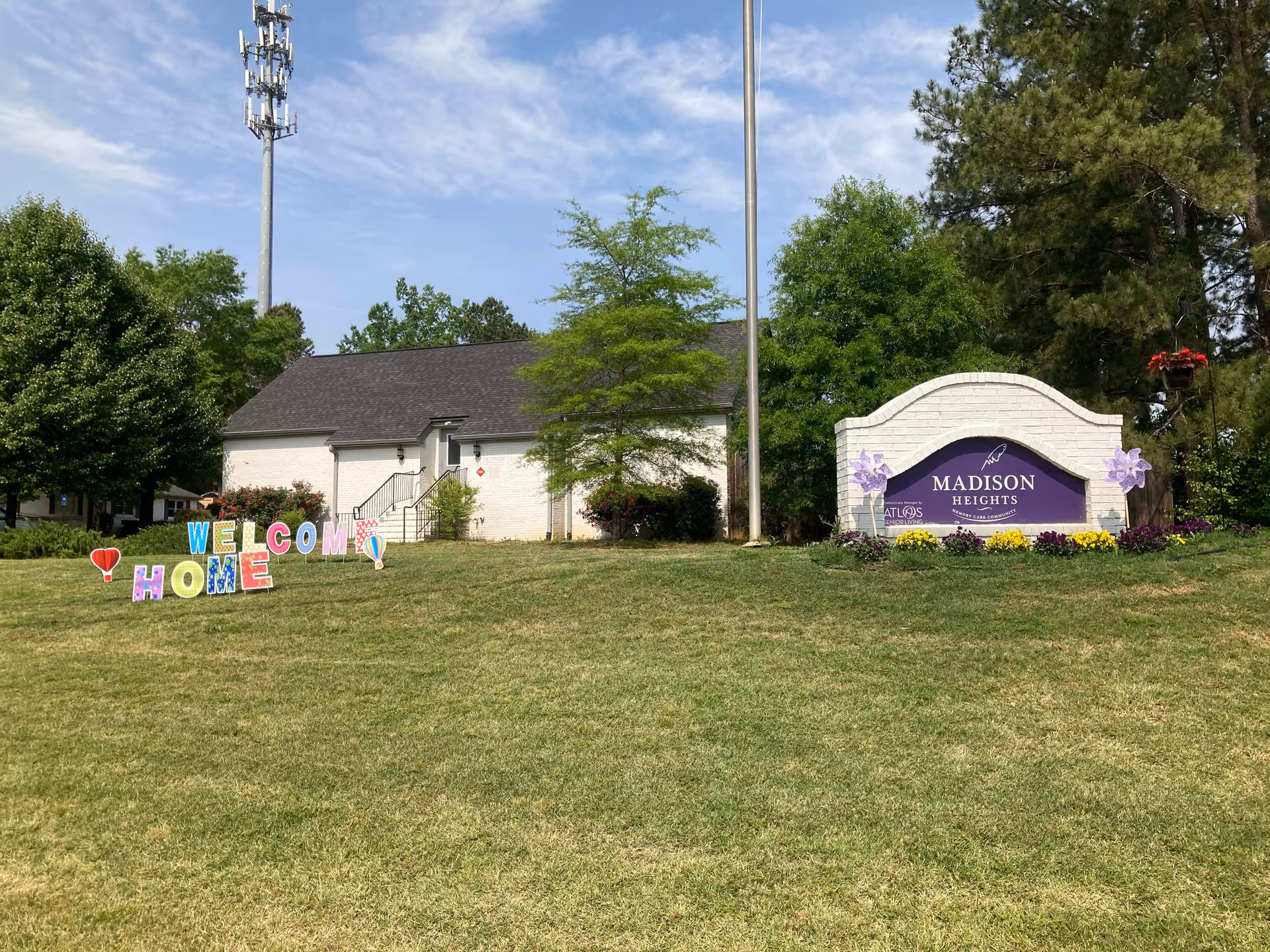 Outdoor view of Madison Heights Evans facility with a white brick sign displaying the facility name surrounded by flowers. A colorful 'Welcome Home' sign is placed on the grass in front of a small white building with a dark roof, trees, and a clear sky in the background.