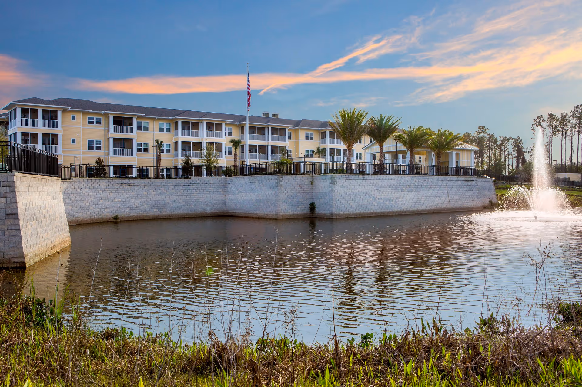 Three-story yellow building by a pond with a fountain, palm trees, and an American flag at sunset.