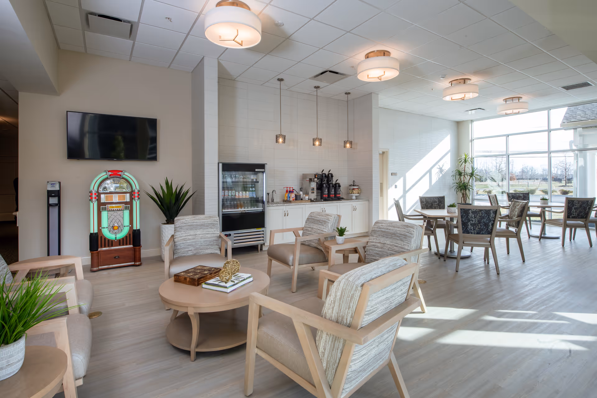 A bright and modern common area with light wood flooring and large windows letting in natural light. The room features several cushioned armchairs arranged around a round coffee table with decorative items, a jukebox, a wall-mounted TV, and a beverage station with coffee dispensers and a refrigerated display case. There are also dining tables and chairs near the windows, and potted plants add greenery to the space.