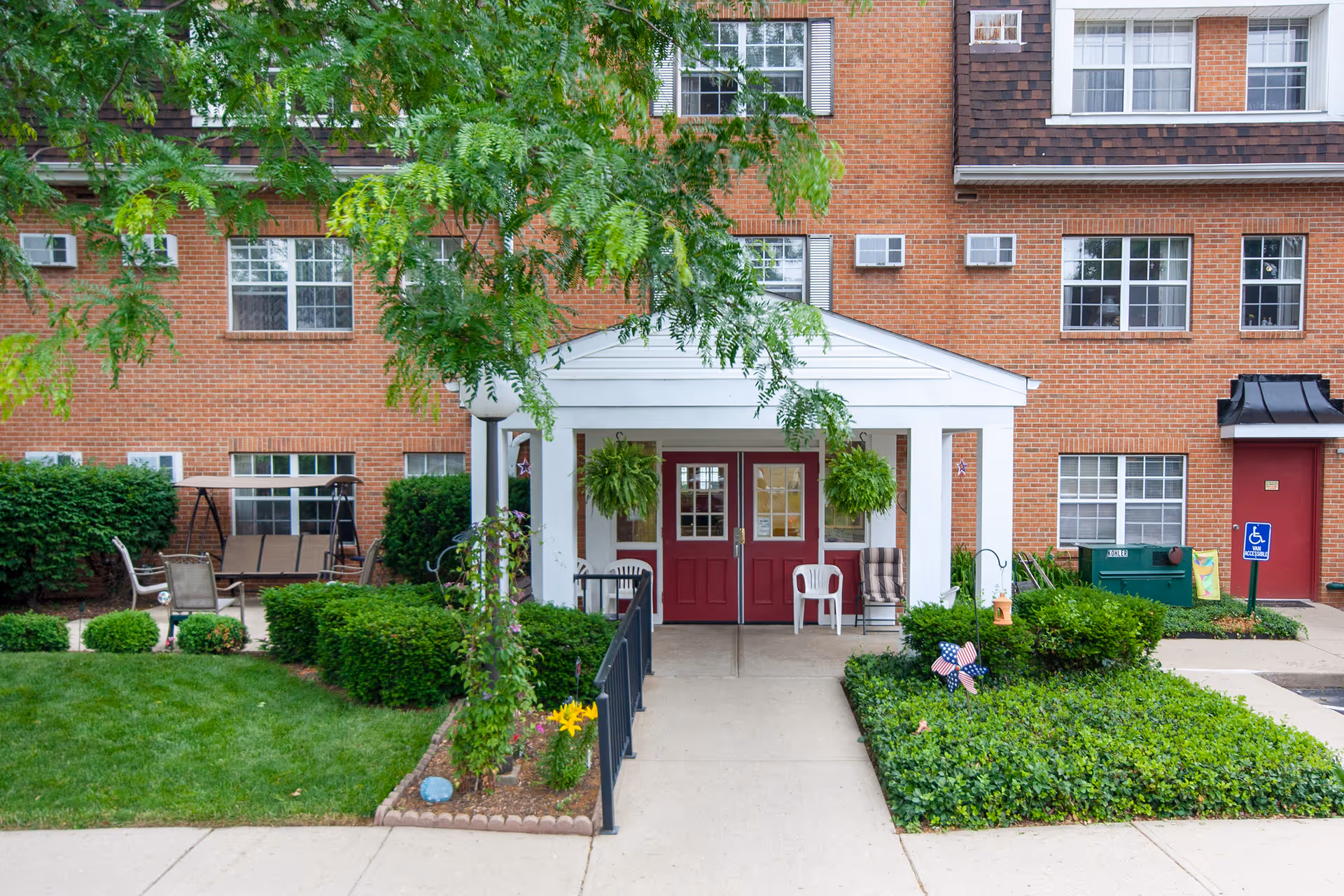 Brick senior living building entrance with a white portico, red double doors, hanging plants and a landscaped walkway.
