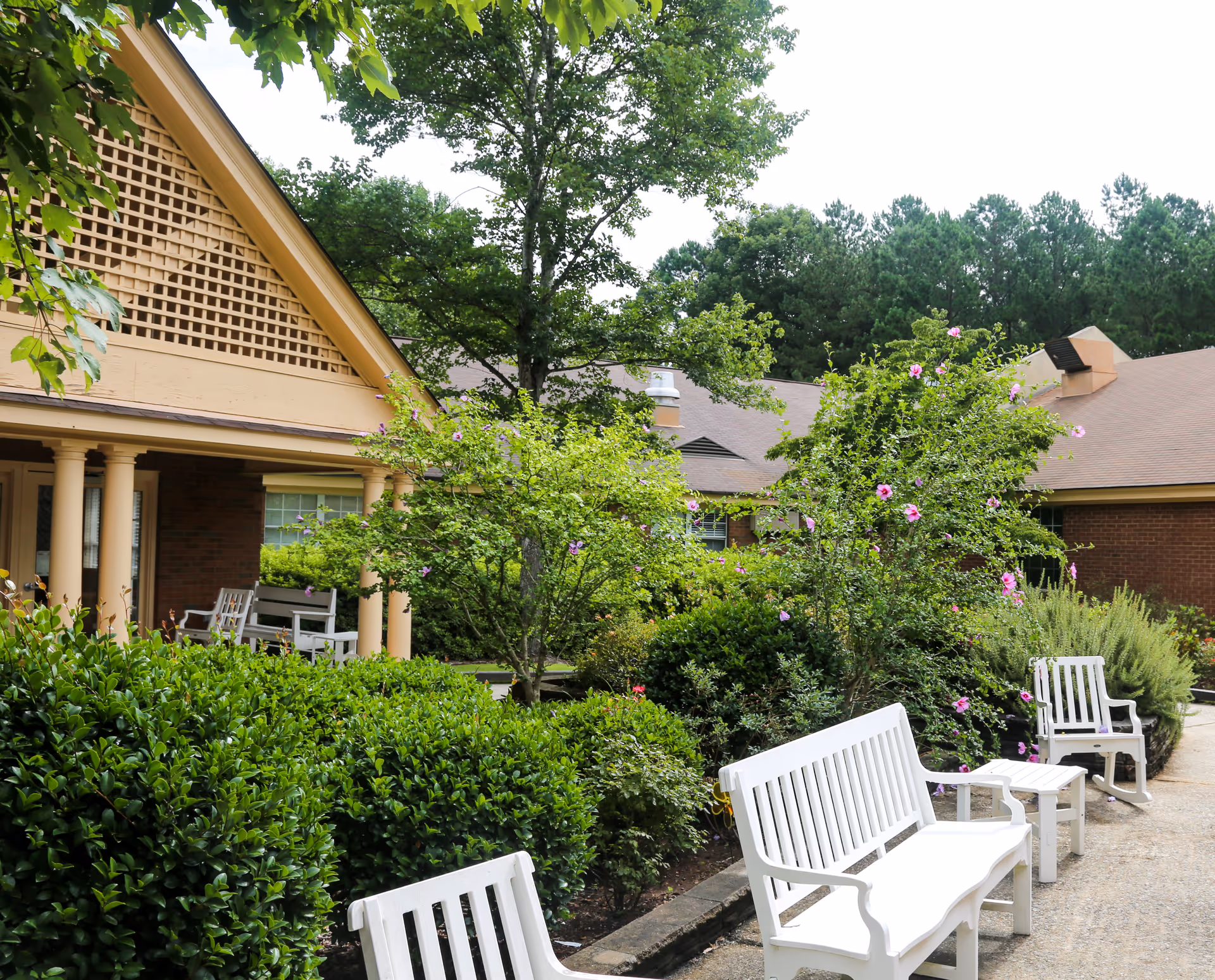 Outdoor seating area at Southland Health & Rehabilitation featuring white benches and chairs along a paved pathway surrounded by green bushes, flowering plants, and trees with a building in the background.