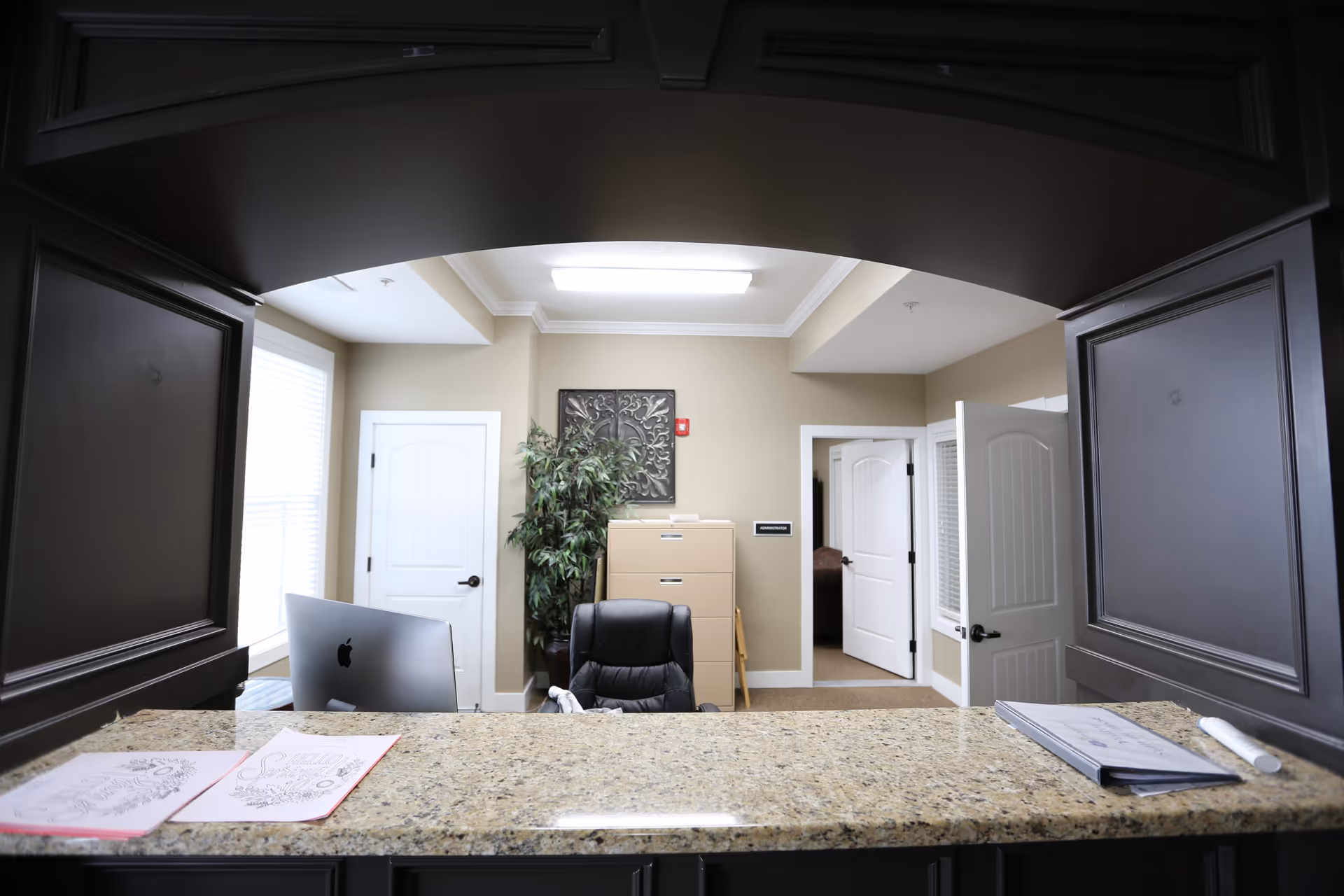 Reception desk area with a granite countertop, a computer, office chair, filing cabinet, potted plant, and open doors leading to other rooms in a well-lit office space.