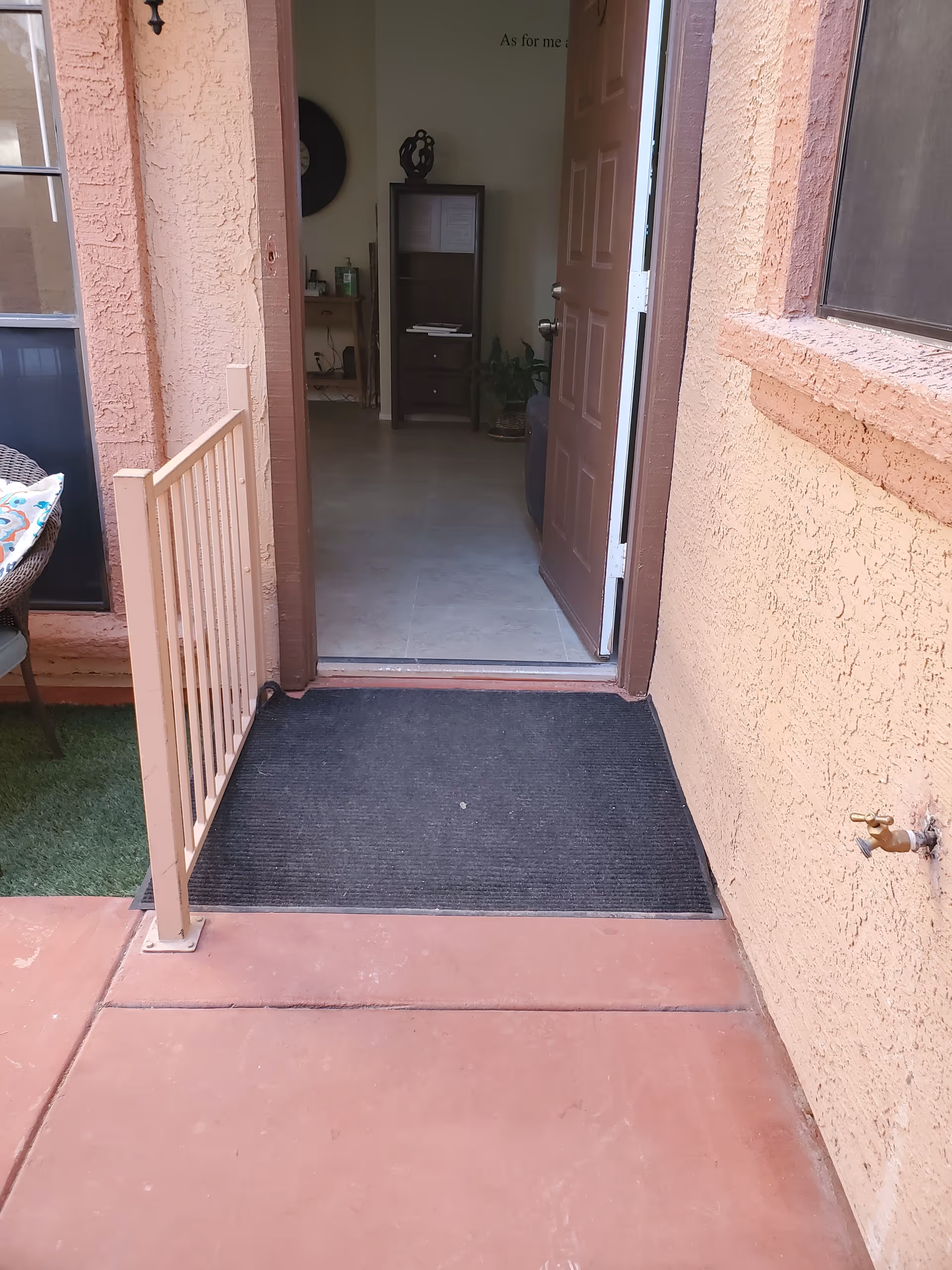 View of an open doorway leading from an outdoor patio area with reddish concrete flooring and a black doormat into an interior room with tiled flooring. The door is brown and partially open. To the left of the doorway is a small beige safety gate and a chair with a colorful cushion. On the right exterior wall, there is a water faucet. Inside, a tall dark wooden shelf and a small table with items on it are visible against the far wall.