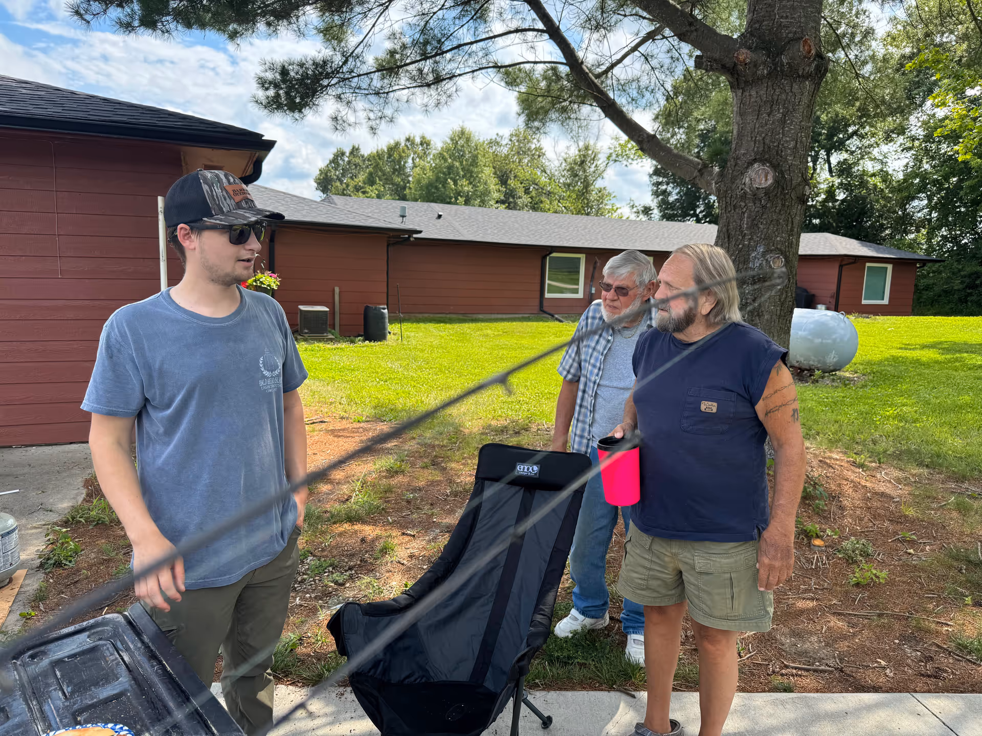 Three men standing outside near a red building with green grass and trees in the background. One man is wearing a blue t-shirt, sunglasses, and a cap, another older man is wearing a plaid shirt and glasses, and the third man is wearing a sleeveless navy shirt and holding a pink cup. There is a black folding chair and a propane tank nearby.